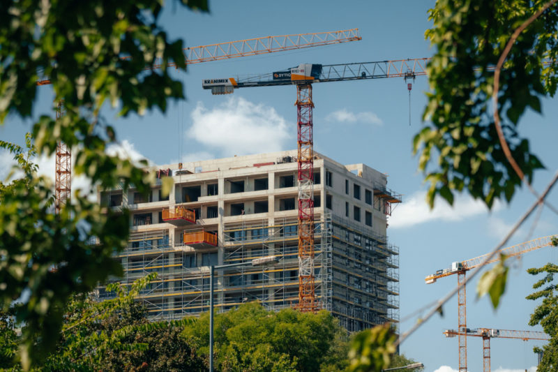 Im Bau befindliches Hochhaus mit Kränen, durch üppig grüne Bäume gesehen.